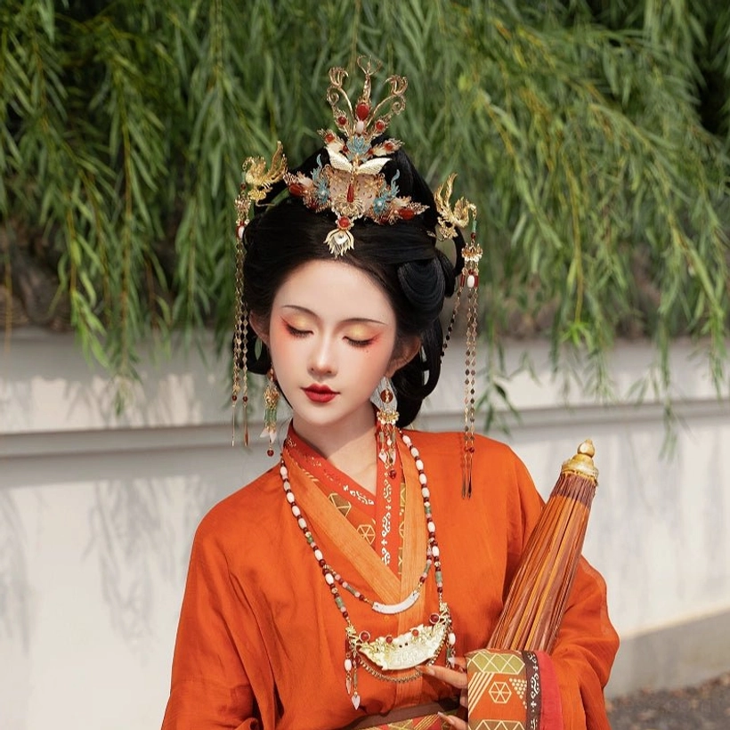 Woman wearing a full hanfu hair styling accessory set with a phoenix crown, traditional updo, hanging ornaments, jade-accented bead details, and an orange robe against willow branches