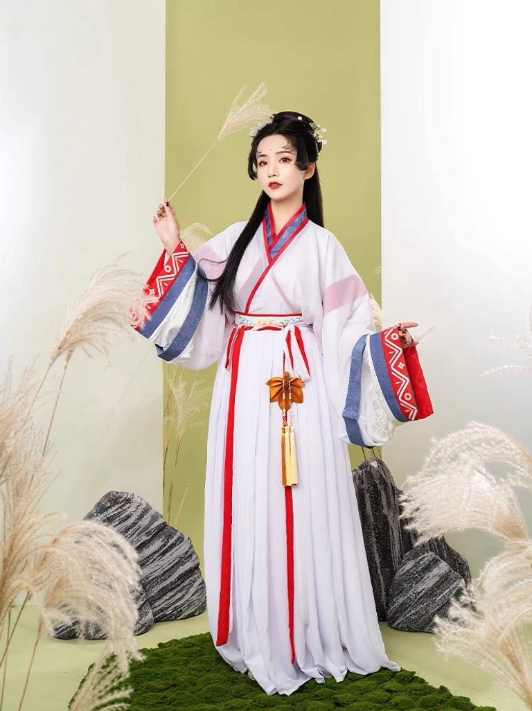 Female hanfu model wearing a white Wei Jin xianxia hanfu with wide sleeves, red and blue geometric cuffs, red waist sash, tassel pendant, and flowing pleated skirt