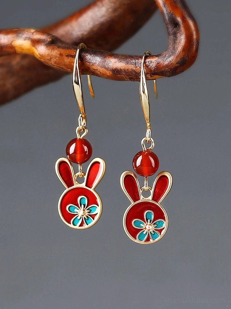 Festive red rabbit drop earrings with gold hooks, red agate beads, and blue floral enamel details hanging from a curved wooden branch