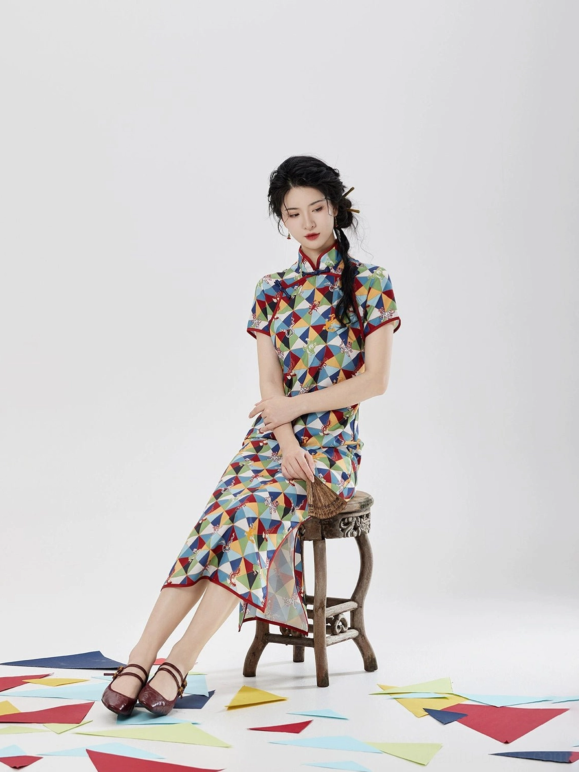 Five Poisons modern Hanfu qipao dress shown in a three-quarter seated view with short sleeves, red stand collar trim, colorful geometric print, and side slit