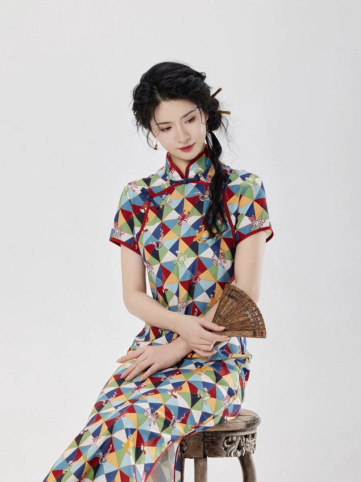 Front seated close-up of Five Poisons patterned cheongsam dress displaying the colorful geometric fabric, red collar piping, and short sleeves