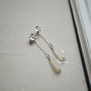 Elegant floral Chinese earrings with polished flower studs, fine silver chains, and translucent off-white teardrop pendants arranged on beige linen fabric