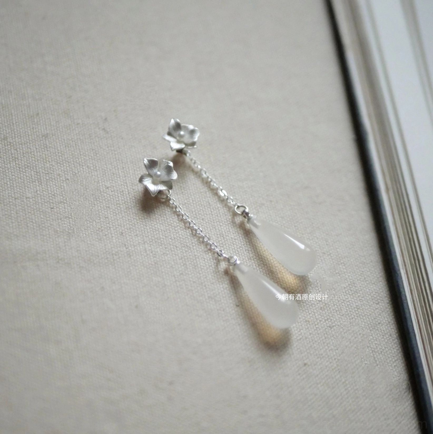 Elegant floral Chinese earrings with polished flower studs, fine silver chains, and translucent off-white teardrop pendants arranged on beige linen fabric