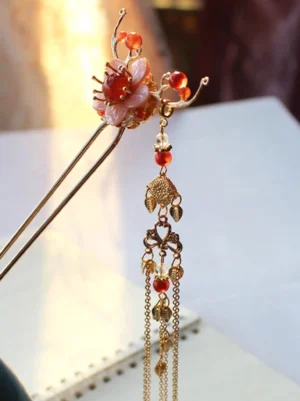 Floral Chinese hair stick with layered pink shell flower, bright red center bead, gold metal details, bow accents, and hanging leaf-shaped tassel
