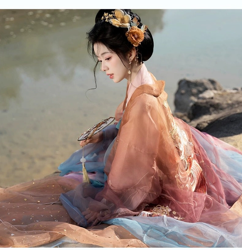 Side sitting view of a woman in romantic Tang Dynasty Hanfu by the riverside with a folding fan and elegant golden flower hair ornaments