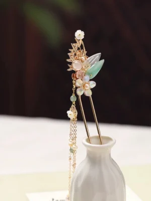 Traditional Chinese hair stick with gold-tone floral ornament, jade-green leaves, pink and white flowers, imitation pearl beads, and long double tassels displayed in a white ceramic vase