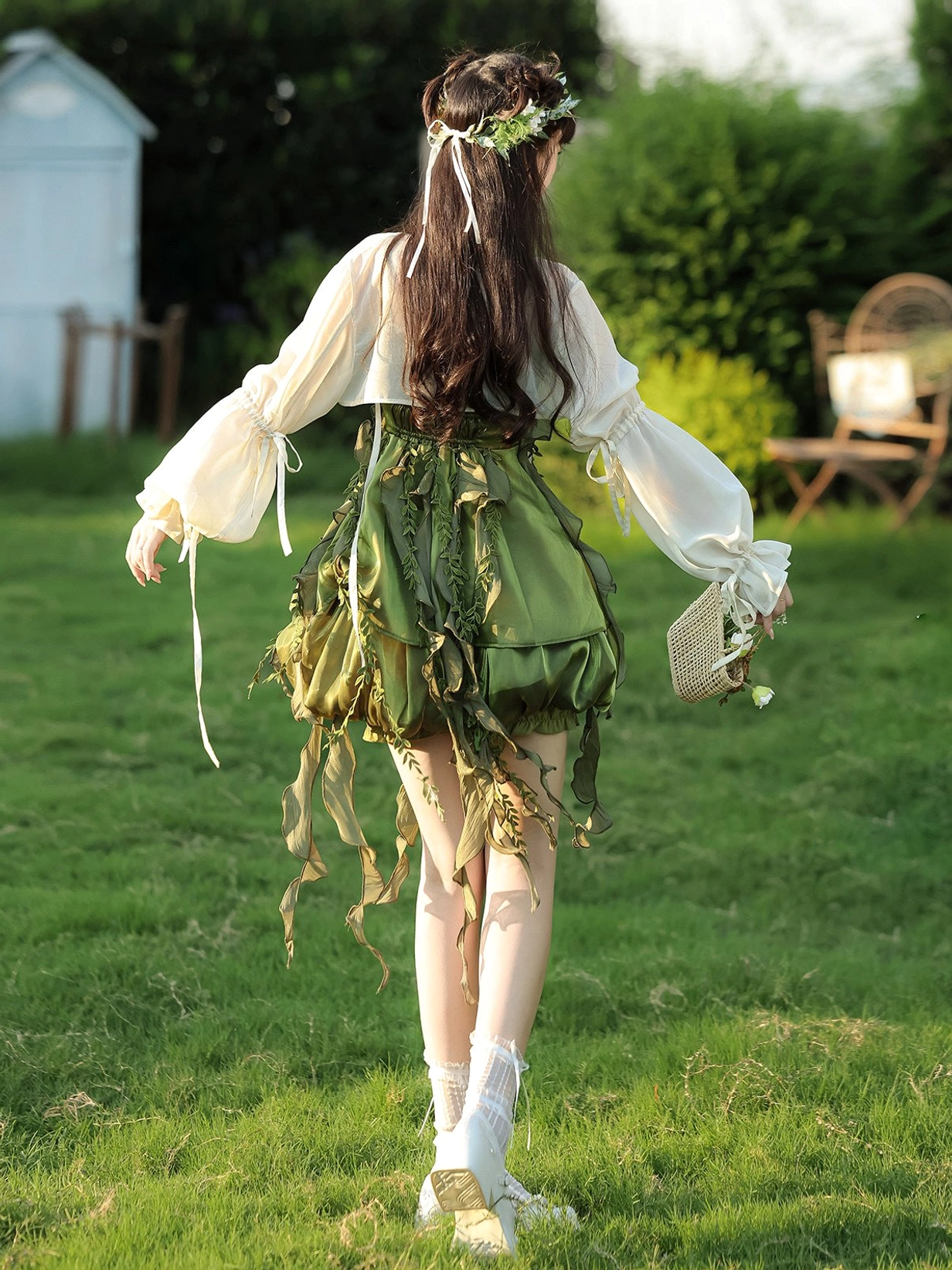 Rear view of forest green forest fairy slip dress and off white cardigan showing leaf tassel decorations with straw handbag styling