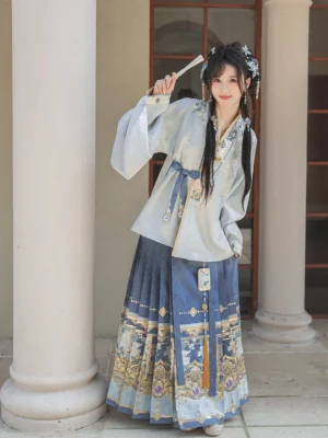 Front view of a traditional Ming Dynasty Hanfu outfit with embroidered cross-collar top, pleated blue mamian skirt, gold floral hem, and matching waist sash