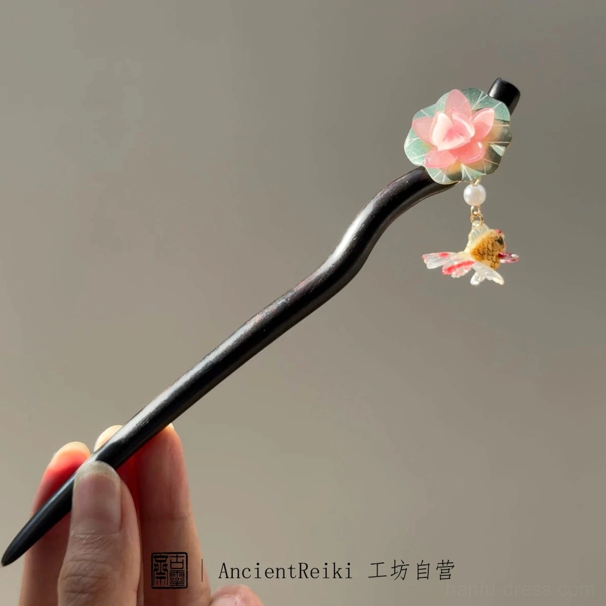 Full-length traditional Chinese koi hair stick held by hand, showing the curved black wooden pin, pink lotus flower, and goldfish ornament