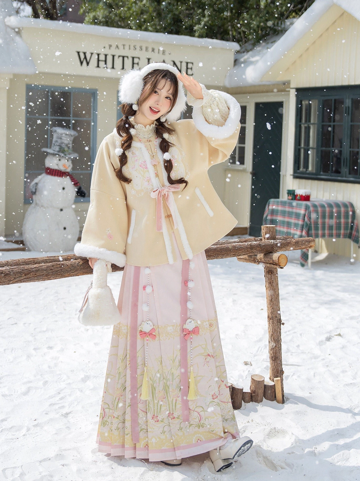 Full length winter Ming style Hanfu costume with beige fur trimmed jacket and pink embroidered Mamian skirt photographed beside a yellow cottage