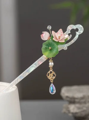 Glazed lotus Chinese hair stick displayed on a white stand, featuring pink lotus flowers, green leaves, an iridescent clear shaft, crystal accents, and a dangling traditional pendant