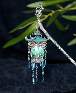 Glowing teal Chinese lantern hair stick with silver carved ornament and blue crystal tassels styled against dark fabric and green leaves for Hanfu hairstyle inspiration