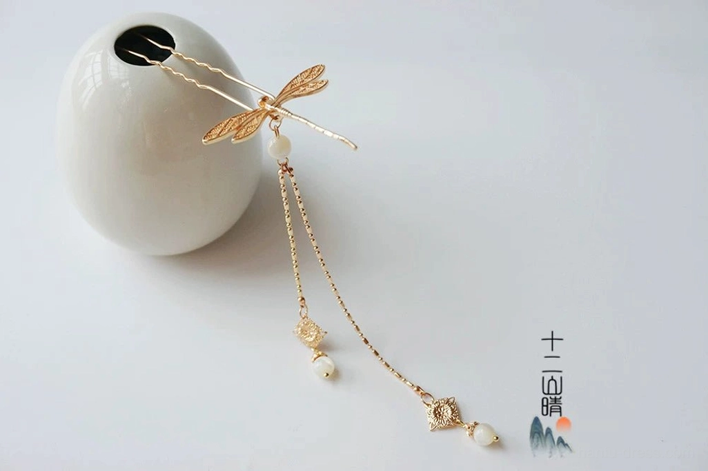 Gold toned Chinese dragonfly tassel hair pin with a white decorative bead and elegant dangling chain pendants beside a white ceramic vase