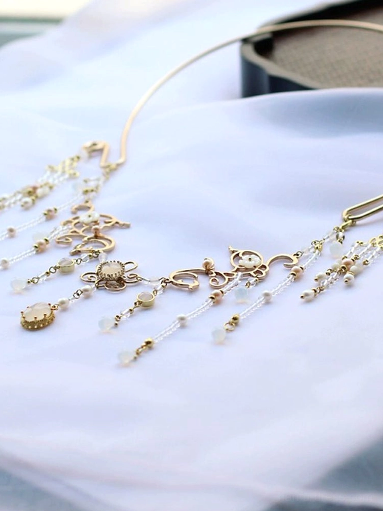 Gold Chinese necklace with compact beaded tassels, floral filigree motifs, and oval gold-toned pendants displayed on white sheer fabric