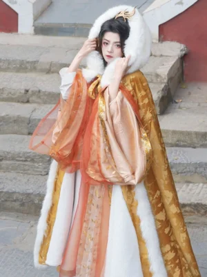 Front three-quarter view of a gold embossed velvet Hanfu cloak with white faux fur hood trim, worn over a traditional Chinese costume in an ancient courtyard