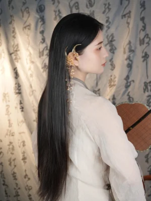 Gold-tone phoenix tassel ear cuff worn on right ear with white Hanfu, long black hair, wooden round fan, and traditional Chinese calligraphy background