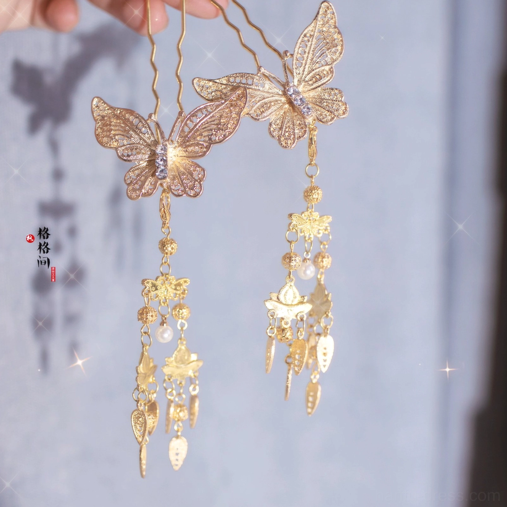 Oblique view of paired golden butterfly tassel hair pins with hollow filigree wings, pearl-like beads, and ornate pendants on a softly sparkling background