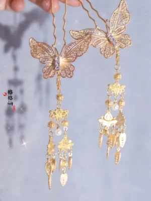Pair of golden butterfly tassel hair pins with openwork filigree wings, sparkling center detail, pearl-like beads, and ornate dangling charms on a light background