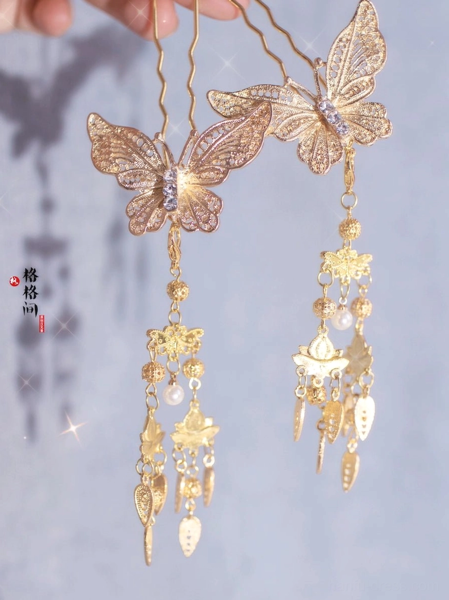 Pair of golden butterfly tassel hair pins with openwork filigree wings, sparkling center detail, pearl-like beads, and ornate dangling charms on a light background