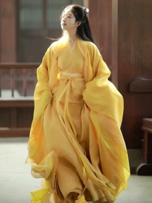 Three-quarter front view of a model wearing a golden yellow Wei Jin Dynasty hanfu robe with a sheer cross-collar design, wide flowing sleeves, tied waist sash, and floor-length skirt in a sunlit traditional Chinese courtyard