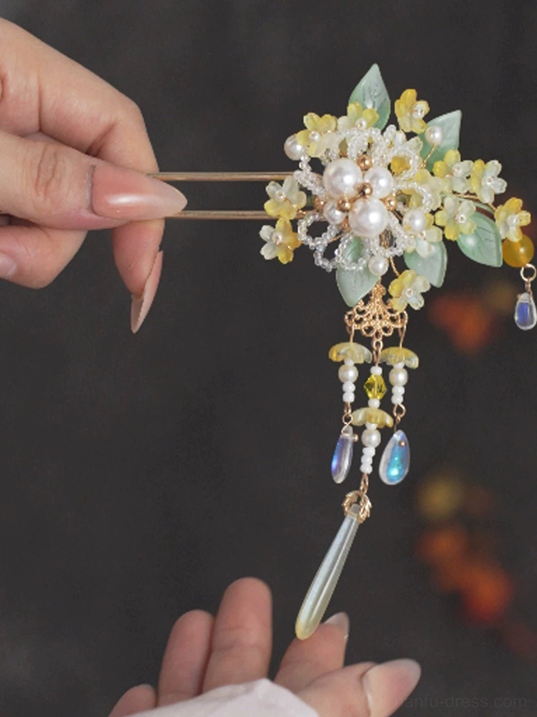 Handheld close-up of golden osmanthus tassel Chinese hair stick showing double-pronged pin floral beads and dangling clear pendants
