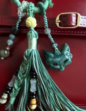 Close-up of green agate fox spirit bag charm Chinese keychain with yellow lotus bead, braided tassel and decorative beads attached to burgundy leather handbag