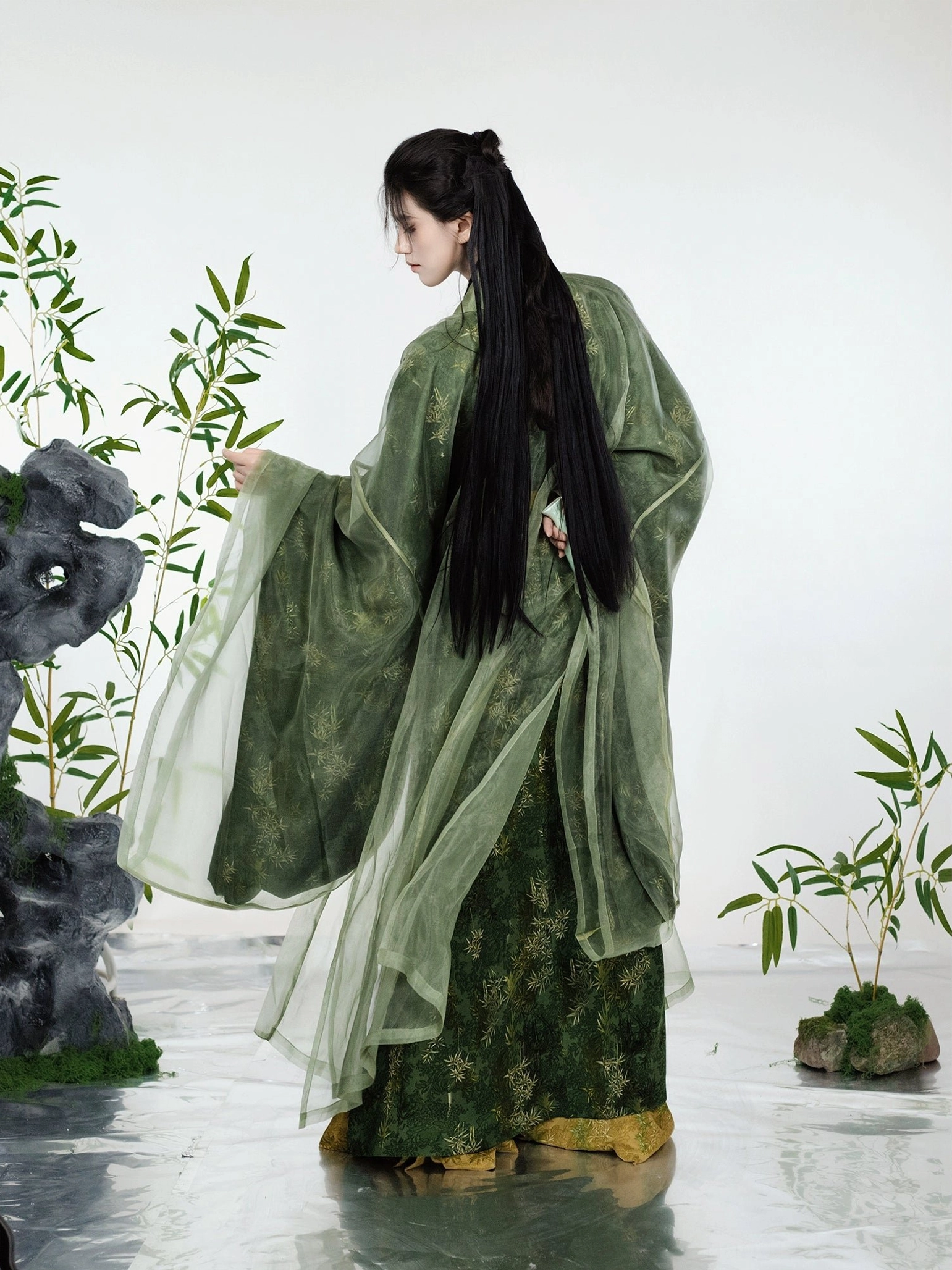 Back-side view of a layered green bamboo leaf print Hanfu costume showing the flowing sheer outer robe, wide sleeves, traditional hairstyle, and golden inner robe trim