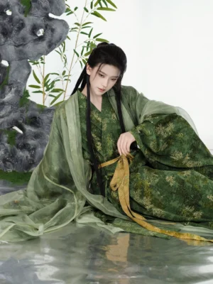 Female model wearing a green bamboo leaf print Song Dynasty Hanfu beizi robe and pants set with wide sheer sleeves, golden sash, traditional hairstyle, rockery and bamboo studio backdrop