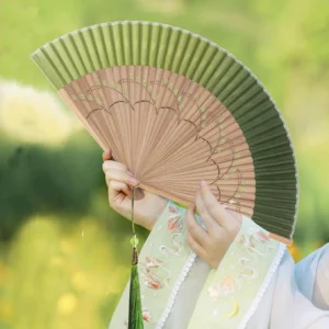 Green Chinese folding hand fan with gradient pleated fabric, carved floral wooden ribs, green tassel, and glass bead held with embroidered Hanfu sleeves
