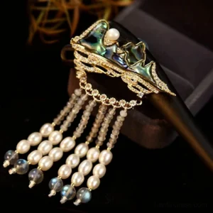 Top-down view of a handmade Chinese hair stick featuring a green enamel mountain charm, pearl centerpiece, sparkling trim, and layered gray-blue pearl tassels beside a jewelry box