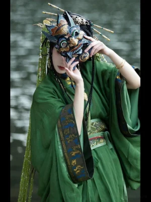 Green Han Dynasty Hanfu straight hem robe front three quarter close up beside the water, showing layered waist sash, flowing sleeves, long tassel hair ornaments, and traditional folk mask styling