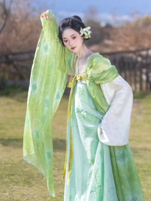 Three-quarter front view of a woman wearing a green Tang Dynasty hanfu set with a short jacket, flowing wrap dress, yellow ribbons, and sheer shawl in a pastoral field