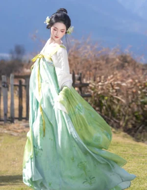 Side view of a flowing green Tang style hanfu wrap dress with ivory sleeves, yellow waist ribbons, and coordinated sheer shawl against a mountain landscape