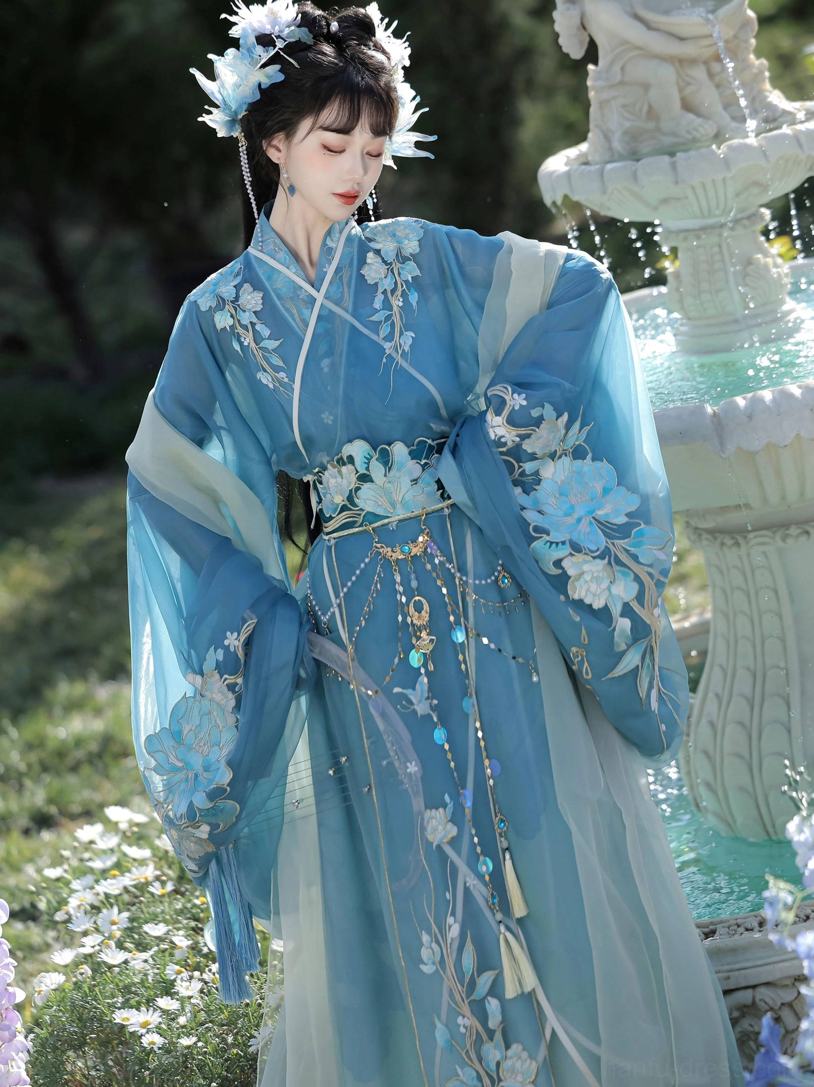 Traditional Chinese Hanfu robe with cross collar, light cyan sheer sleeves, floral embroidery, and jeweled waist accessories shown in a front garden portrait