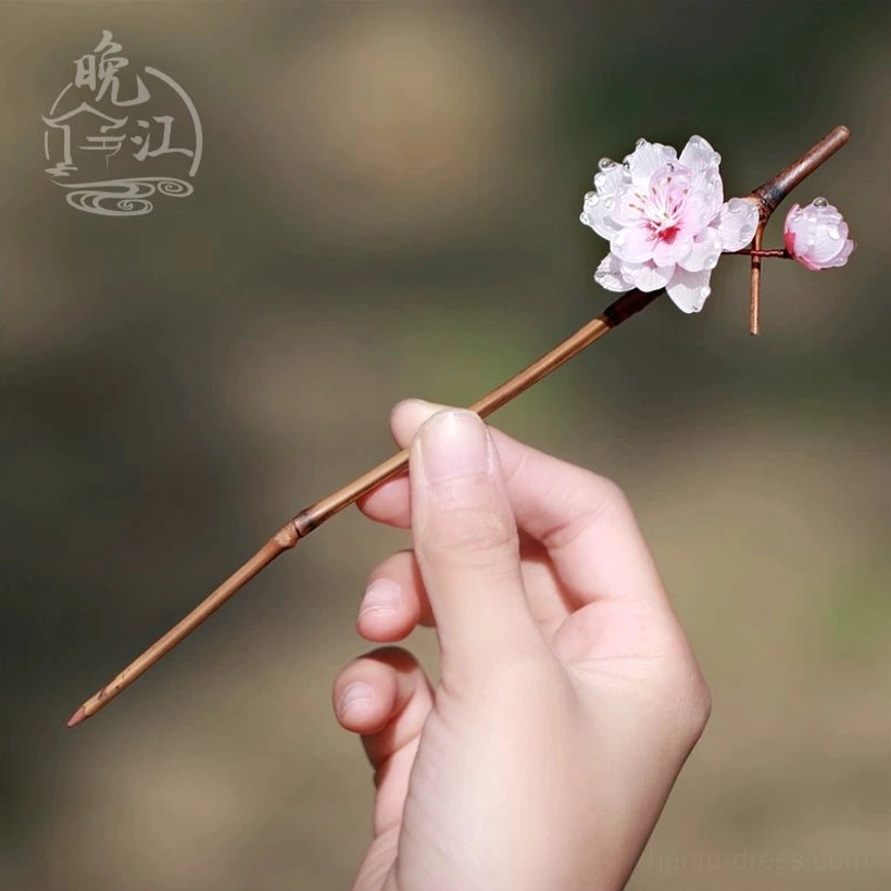 Handheld Chinese plum blossom hair stick with a small pink flower tiny bud and slender brown wooden shaft styled against a soft outdoor background