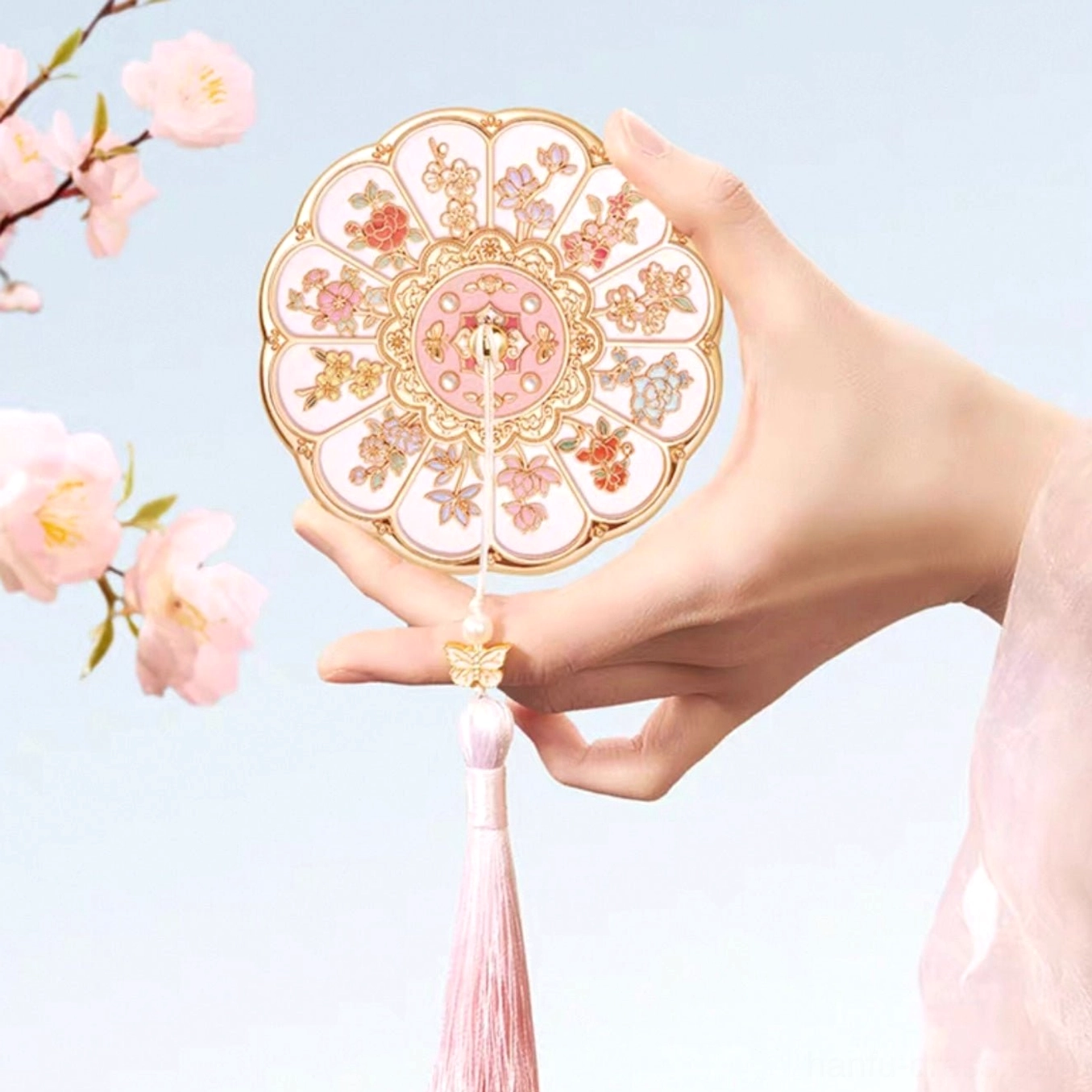 Handheld Chinese pocket mirror with intricate floral cover, long pink tassel, and gold butterfly charm under cherry blossoms
