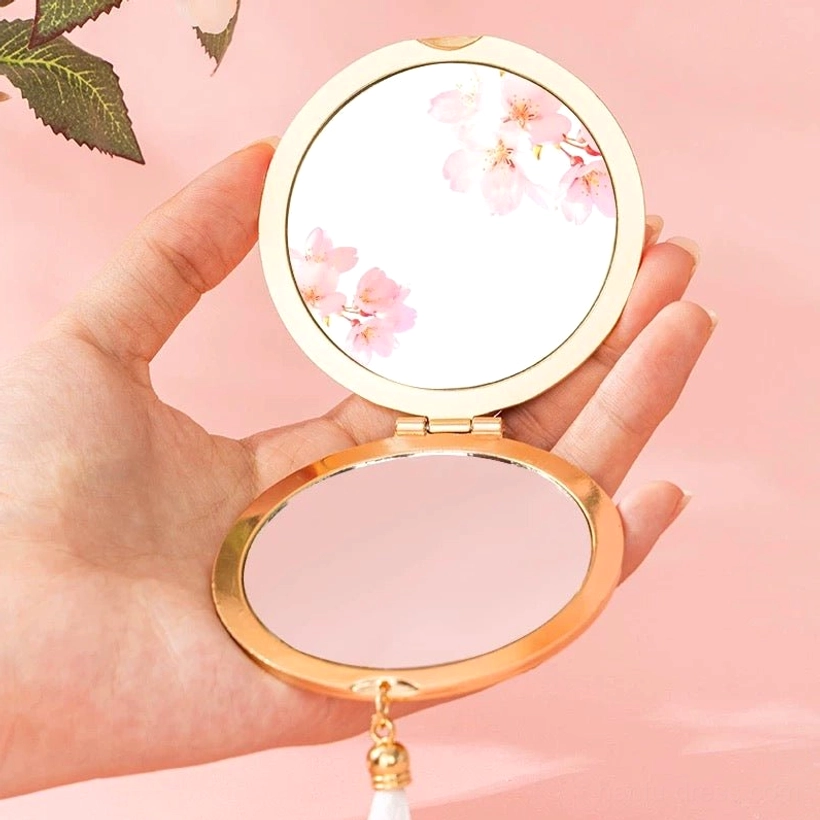 Handheld Chinese pocket mirror opened to show the reflective surface, gold-tone frame, floral artwork, and white tassel pendant