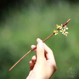 Hand holding a traditional osmanthus flower Chinese hair stick with delicate yellow blossoms, ivory floral details, and brown wooden shaft outdoors