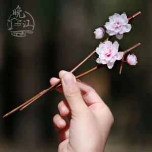 Handheld pink plum blossom Chinese hair stick with layered floral petals small flower buds and brown wooden shaft against a natural background