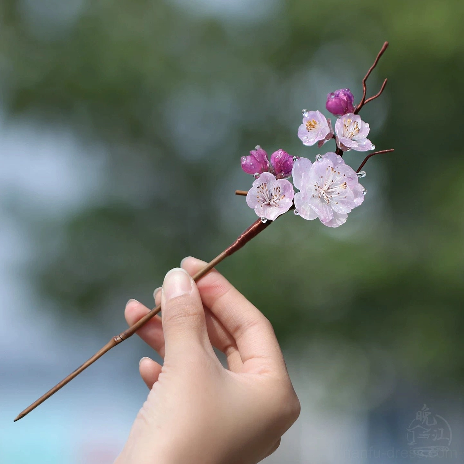 Hand holding a traditional Chinese plum blossom hair stick with pink flowers and purple buds against a blurred natural green background