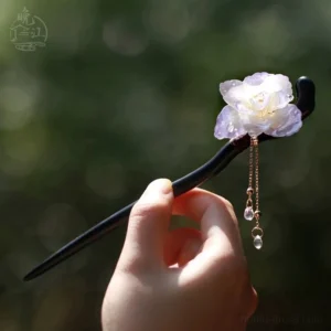 Hand holding a traditional Chinese flower hair stick with translucent purple resin petals, golden tassels, and water-drop crystal pendants