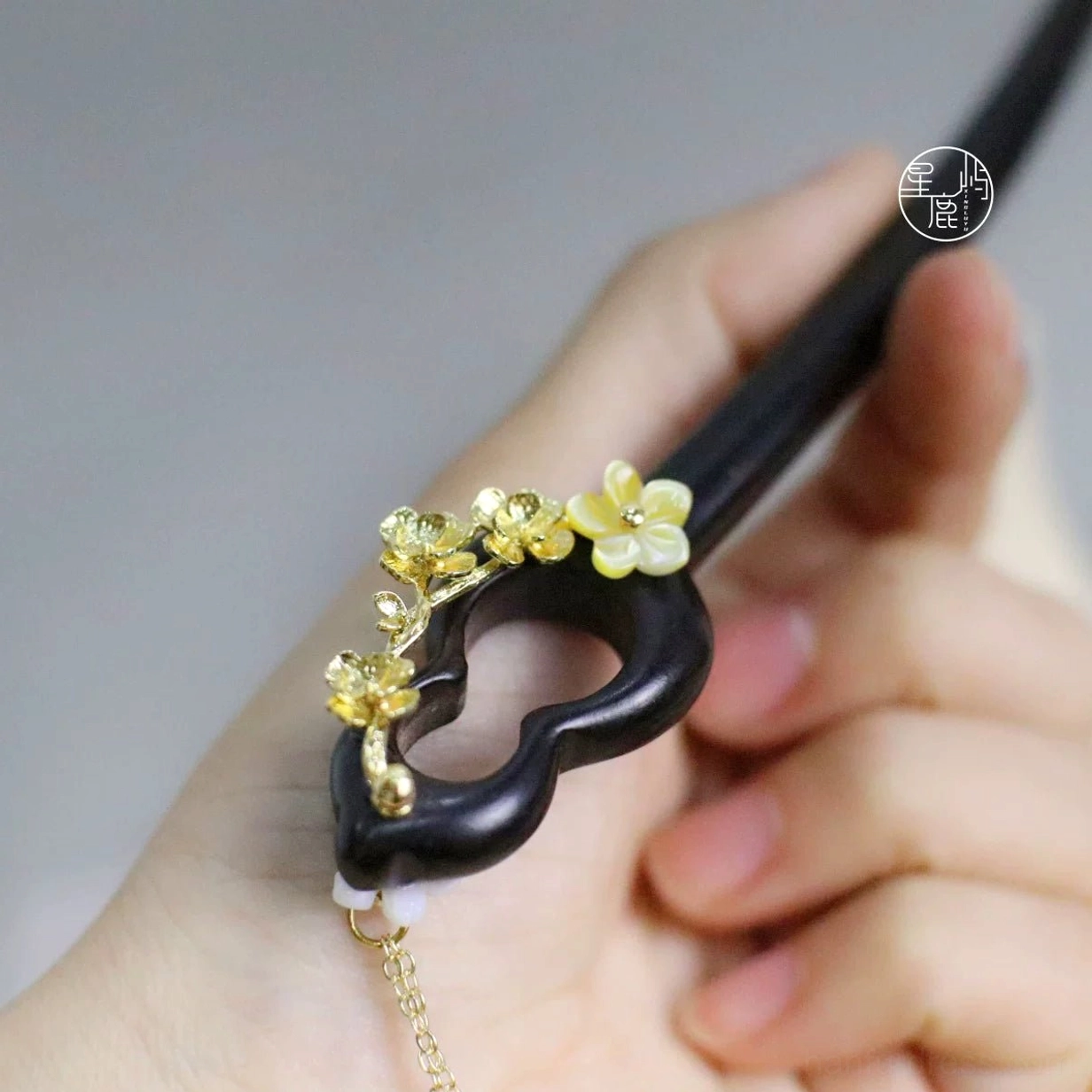 Handheld close-up of a traditional Chinese dark wood hair stick with gourd-shaped carved ornament golden flowers and delicate hanging tassel chain