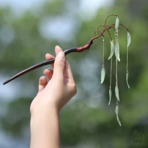 Hand holding a traditional Chinese willow hair stick with elegant jade-green leaf pendants and delicate gold-toned tassel chains in sunlit outdoor greenery