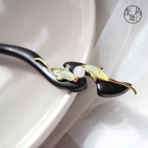 Close-up of handmade Chinese hair stick with curved black wood shaft, paired gold fish ornaments, green accents, and central white decorative bead
