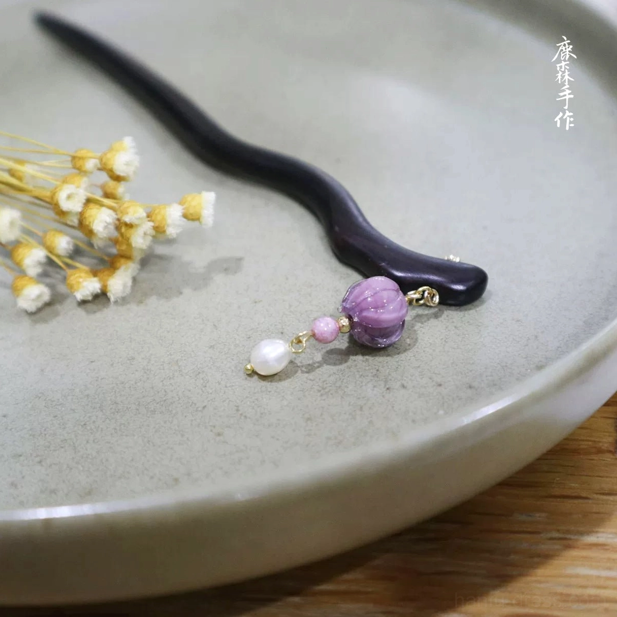 Handmade ebony Chinese hair stick with wavy dark wood design, purple pumpkin bead, pink bead, white pearl pendant, and gold-tone details displayed with dried flowers
