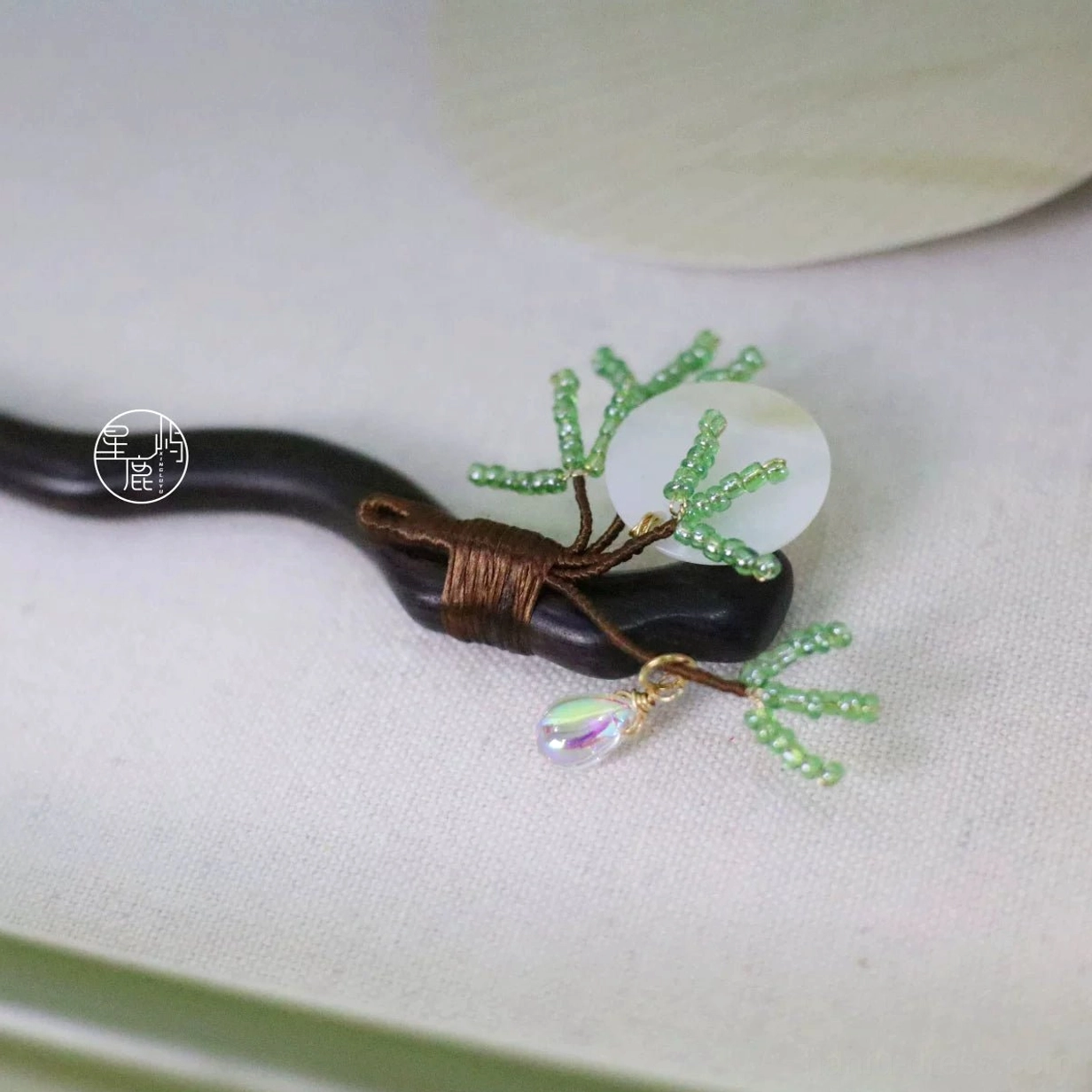 Close-up top view of a handmade pine and moon Hanfu hair accessory on beige fabric, showing textured dark wood, green beaded pine twigs, round shell ornament, and iridescent waterdrop