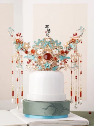 Large traditional Chinese hanfu bridal hair crown with turquoise cloisonne-style floral carvings, central red gemstone accent, white flowers, red beads, pearl pendants, and long dangling tassels displayed on a ceramic stand