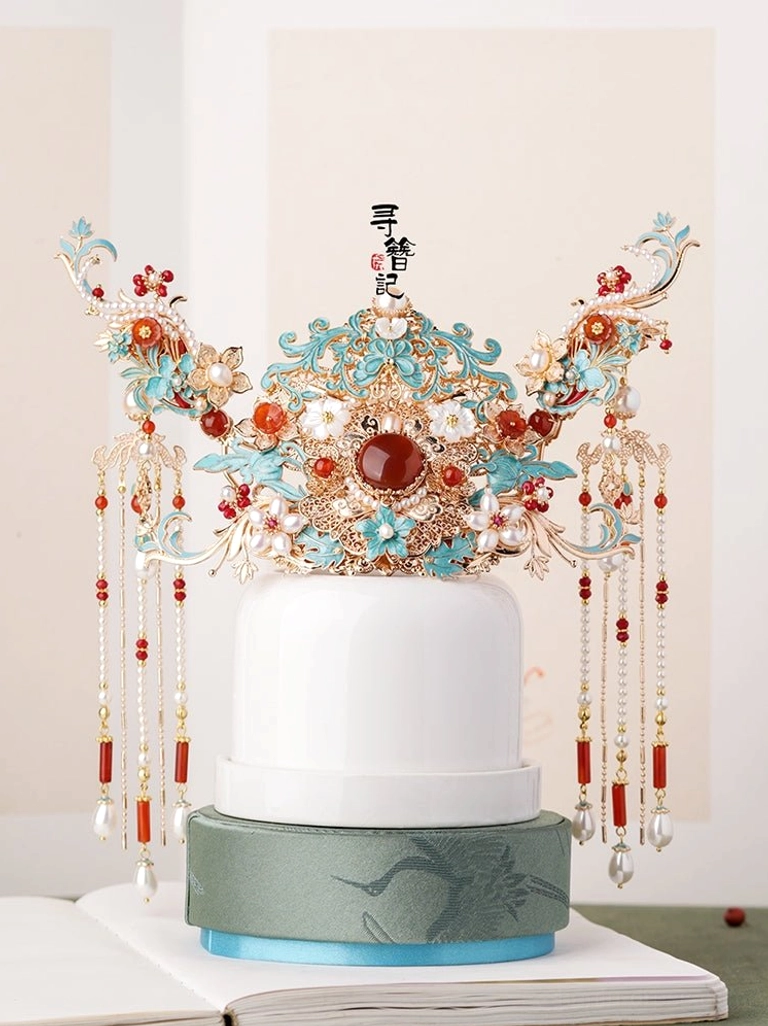 Large traditional Chinese hanfu bridal hair crown with turquoise cloisonne-style floral carvings, central red gemstone accent, white flowers, red beads, pearl pendants, and long dangling tassels displayed on a ceramic stand
