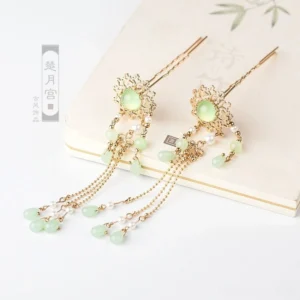 Chinese Hanfu hair pin pair with gold-tone flower filigree, jade-green decorative stones, layered chain tassels, and green bead pendants on a vintage poetry notebook
