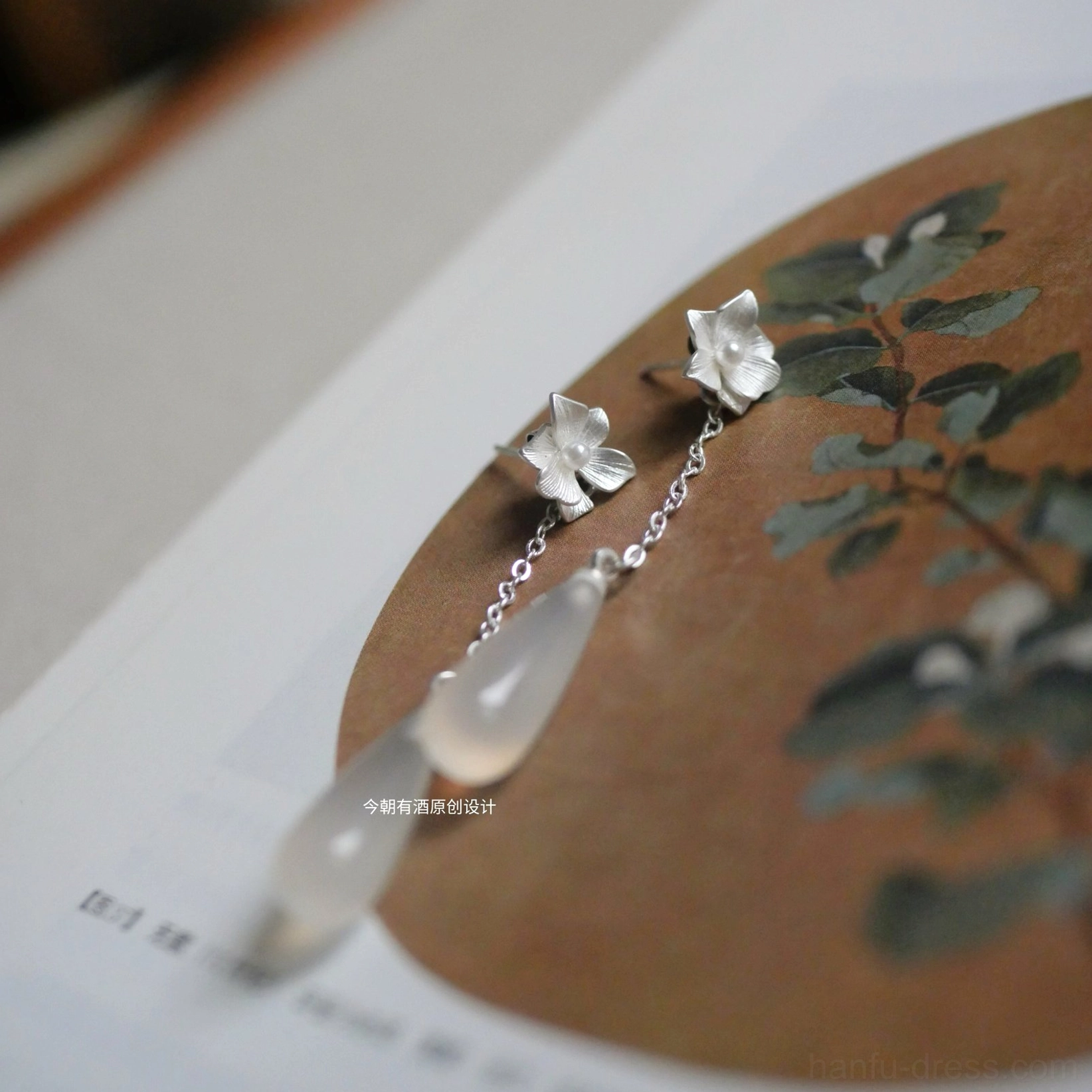 Hanfu floral jade dangle earrings with silver flower ear posts, thin hanging chains, and warm-toned teardrop pendants shown over a traditional Chinese painting